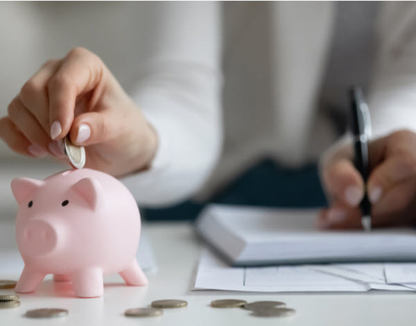 Step Up - Learn helpful money habits Woman putting a coin in a piggy bank while writing on her notebook