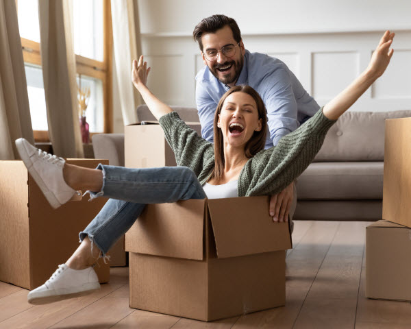Step-Up- First home buyer Full length overjoyed young bearded man in glasses pushing laughing wife in carton box