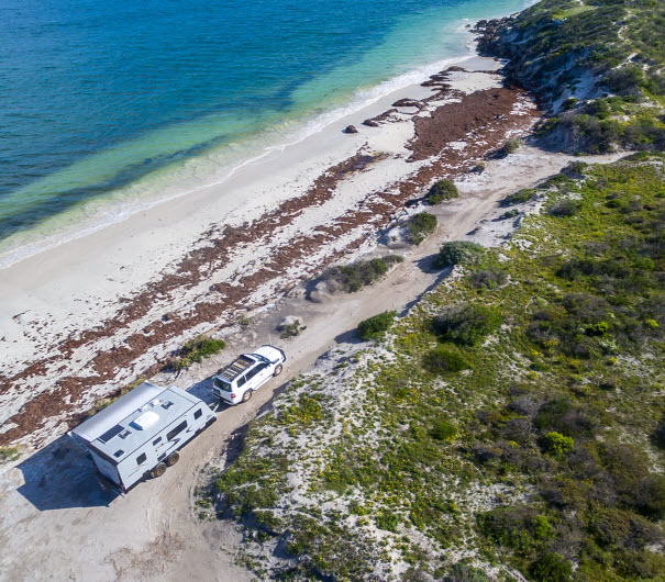Landscape aerial view of 4WD and modern caravan parked adjacent to a sparkling sunny beach